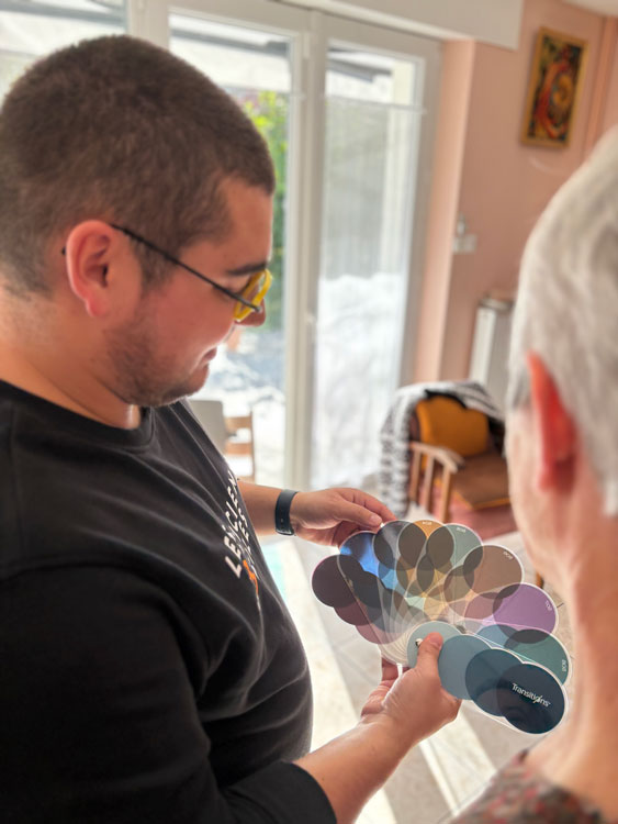 Sur cette photo, à gauche il y a un homme à lunettes jaune. Cet homme est un opticien à domicile, faisant parti du groupe Les Opticiens Mobiles. Sur la droite il y a sa cliente souhaitant changer de lunettes. Au milieu de la photo, un pantonier de colorie de verres Essilor Transition. La cliente choisi la couleur de ses futurs verres photochromiques. 
